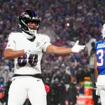 Isaiah Likely von den Baltimore Ravens, in weißer Uniform, gestikuliert mit einem ausgestreckten Arm während eines verschneiten Nachtspiels. In der Nähe steht ein Spieler der Buffalo Bills in blauer Uniform mit der Nummer 31 mit dem Rücken zum Publikum. Die Menge im Stadion ist im Hintergrund verschwommen. Diese Beschreibung wurde mit der FootballR KI automatisch generiert.