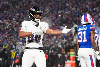 Isaiah Likely von den Baltimore Ravens, in weißer Uniform, gestikuliert mit einem ausgestreckten Arm während eines verschneiten Nachtspiels. In der Nähe steht ein Spieler der Buffalo Bills in blauer Uniform mit der Nummer 31 mit dem Rücken zum Publikum. Die Menge im Stadion ist im Hintergrund verschwommen. Diese Beschreibung wurde mit der FootballR KI automatisch generiert.