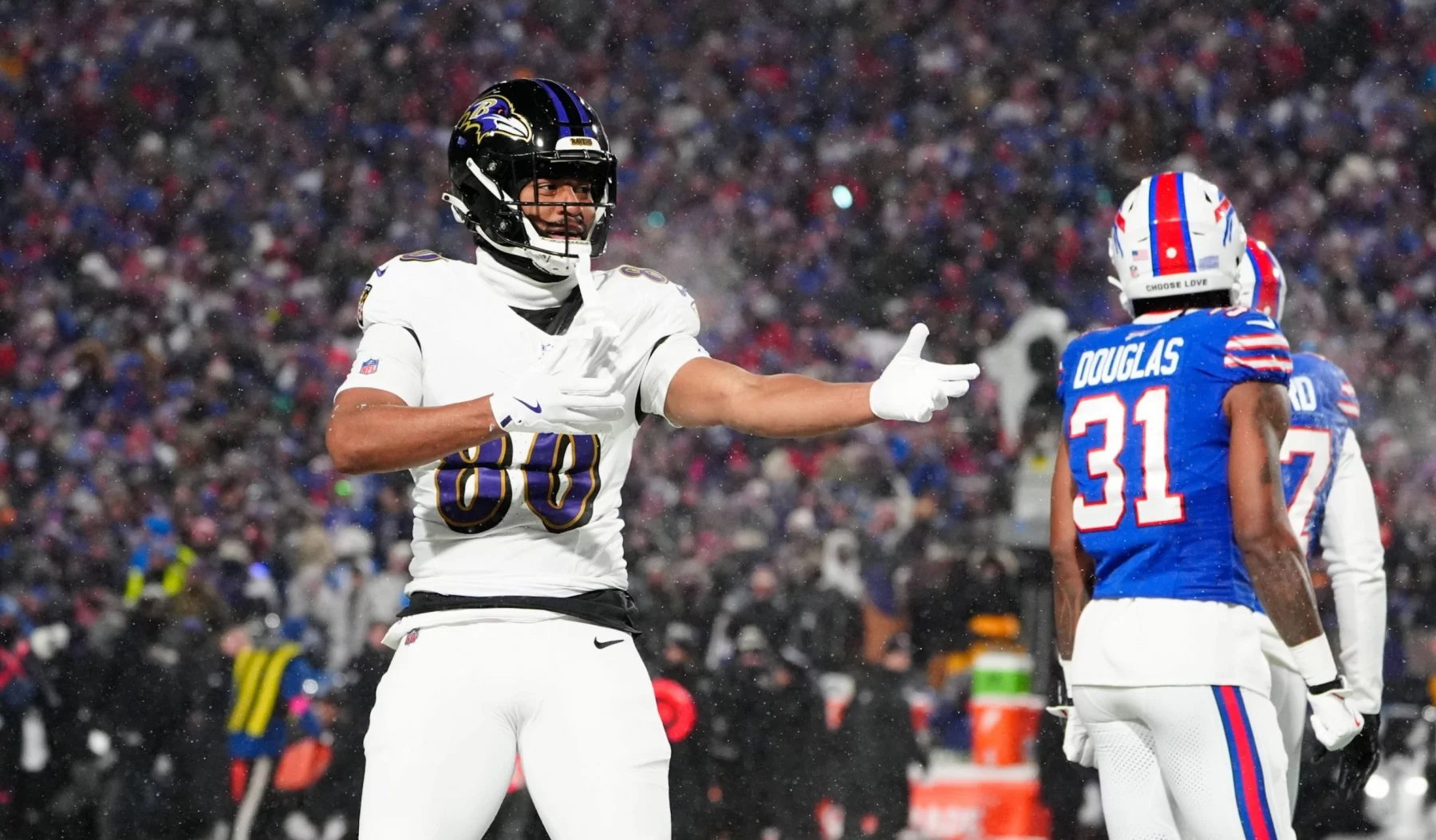 Isaiah Likely von den Baltimore Ravens, in weißer Uniform, gestikuliert mit einem ausgestreckten Arm während eines verschneiten Nachtspiels. In der Nähe steht ein Spieler der Buffalo Bills in blauer Uniform mit der Nummer 31 mit dem Rücken zum Publikum. Die Menge im Stadion ist im Hintergrund verschwommen. Diese Beschreibung wurde mit der FootballR KI automatisch generiert.