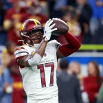 Terry McLaurin, der ein weißes Trikot der Washington Commanders mit der Nummer 17 trägt, fängt während eines Spiels einen Football, während Zuschauer und andere Spieler im Hintergrund verschwimmen. Diese Beschreibung wurde mit der FootballR KI automatisch generiert.