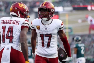 Zwei Footballspieler der Washington Commanders feiern auf dem Spielfeld. Terry McLaurin, Nummer 17, hält einen Ball und ruft der Nummer 14 energisch zu. Beide tragen weiße Trikots mit burgunderroten und goldenen Akzenten, während Spieler der Philadelphia Eagles unscharf zu sehen sind. Diese Beschreibung wurde mit der FootballR KI automatisch generiert.