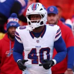 Von Miller in einem weißen Buffalo-Bills-Trikot mit der Nummer 40, mit Helm und Handschuhen, steht auf dem Spielfeld. Inmitten von Gerüchten über Tauschgeschäfte wie "Von Miller wechselt zu den Commanders" erscheinen Teamkollegen und Trainer in Bills-Kleidung im verschwommenen Hintergrund. Diese Beschreibung wurde mit der FootballR KI automatisch generiert.