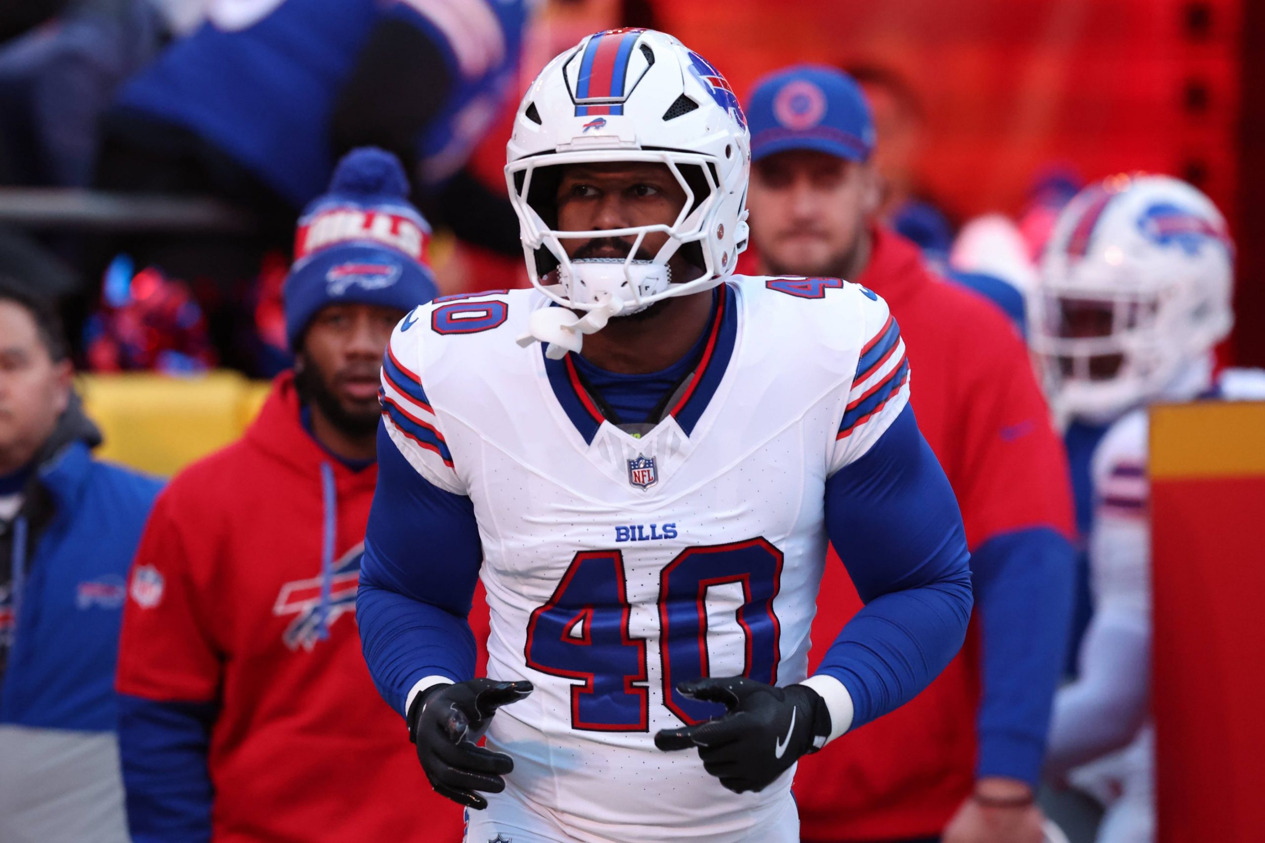 Von Miller in einem weißen Buffalo-Bills-Trikot mit der Nummer 40, mit Helm und Handschuhen, steht auf dem Spielfeld. Inmitten von Gerüchten über Tauschgeschäfte wie "Von Miller wechselt zu den Commanders" erscheinen Teamkollegen und Trainer in Bills-Kleidung im verschwommenen Hintergrund. Diese Beschreibung wurde mit der FootballR KI automatisch generiert.