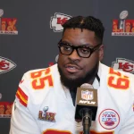 Der Kansas City Chiefs-Spieler Trey Smith, der ein weißes Trikot mit der Nummer 65 trägt, spricht während einer Super Bowl LIV-Presseveranstaltung in ein Mikrofon. Im Hintergrund sind die Logos der Chiefs und des Super Bowls zu sehen. Diese Beschreibung wurde mit der FootballR KI automatisch generiert.