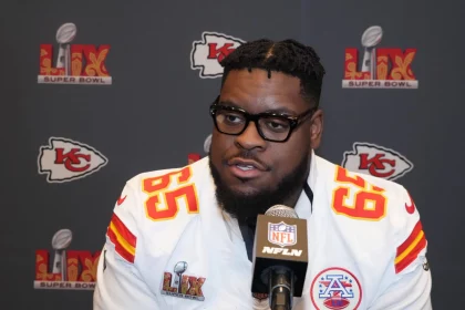 Der Kansas City Chiefs-Spieler Trey Smith, der ein weißes Trikot mit der Nummer 65 trägt, spricht während einer Super Bowl LIV-Presseveranstaltung in ein Mikrofon. Im Hintergrund sind die Logos der Chiefs und des Super Bowls zu sehen. Diese Beschreibung wurde mit der FootballR KI automatisch generiert.