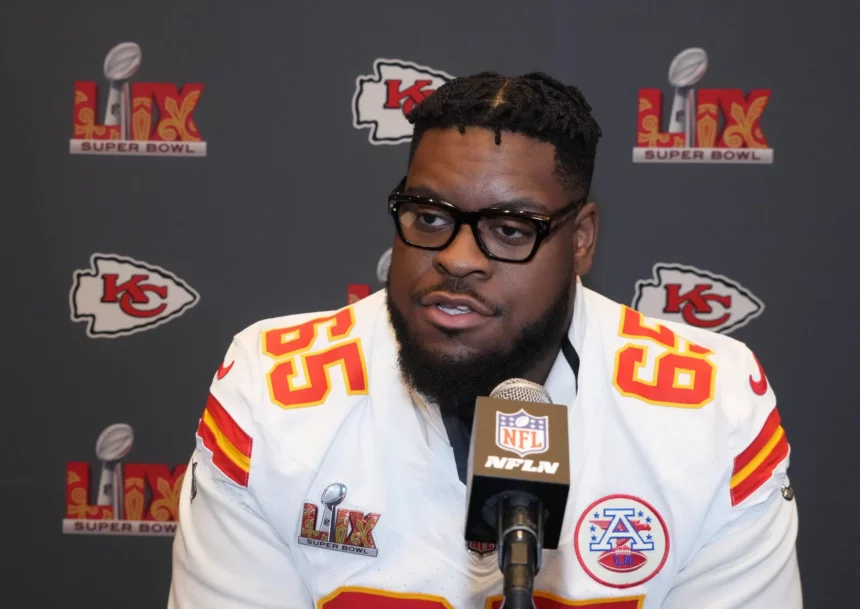 Der Kansas City Chiefs-Spieler Trey Smith, der ein weißes Trikot mit der Nummer 65 trägt, spricht während einer Super Bowl LIV-Presseveranstaltung in ein Mikrofon. Im Hintergrund sind die Logos der Chiefs und des Super Bowls zu sehen. Diese Beschreibung wurde mit der FootballR KI automatisch generiert.