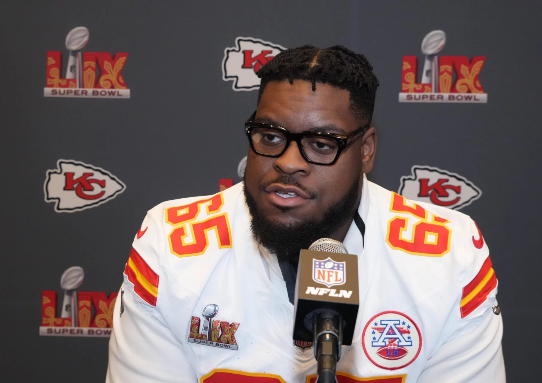 Der Kansas City Chiefs-Spieler Trey Smith, der ein weißes Trikot mit der Nummer 65 trägt, spricht während einer Super Bowl LIV-Presseveranstaltung in ein Mikrofon. Im Hintergrund sind die Logos der Chiefs und des Super Bowls zu sehen. Diese Beschreibung wurde mit der FootballR KI automatisch generiert.