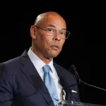 Lloyd Howell mit Brille, dunklem Anzug, weißem Hemd und blauer Krawatte steht an einem Podium und spricht in ein Mikrofon vor einem schwarzen Hintergrund. Er schaut mit ernster Miene leicht zur Seite, als die Nachricht kommt, dass Lloyd Howell zurücktritt. Diese Beschreibung wurde mit der FootballR KI automatisch generiert.