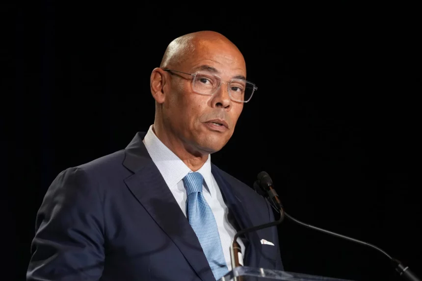 Lloyd Howell mit Brille, dunklem Anzug, weißem Hemd und blauer Krawatte steht an einem Podium und spricht in ein Mikrofon vor einem schwarzen Hintergrund. Er schaut mit ernster Miene leicht zur Seite, als die Nachricht kommt, dass Lloyd Howell zurücktritt. Diese Beschreibung wurde mit der FootballR KI automatisch generiert.