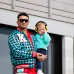 Patrick Mahomes mit Sonnenbrille, rotem Hemd und blauer Jacke mit Hahnentrittmuster hält ein kleines Kind in einem blaugrünen Pullover und gelben Kopfhörern mit Geräuschunterdrückung draußen in der Nähe von Gebäude 106". Beide schauen in die Kamera, vielleicht auf dem Weg zu einer NFL 18 Spiele-Veranstaltung. Das Kind berührt die Kopfhörer. Diese Beschreibung wurde mit der FootballR KI automatisch generiert.