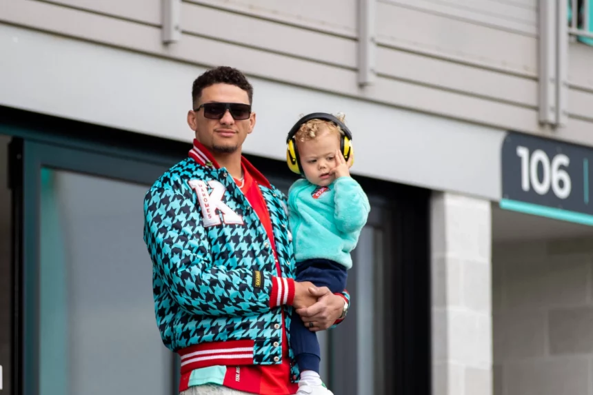 Patrick Mahomes mit Sonnenbrille, rotem Hemd und blauer Jacke mit Hahnentrittmuster hält ein kleines Kind in einem blaugrünen Pullover und gelben Kopfhörern mit Geräuschunterdrückung draußen in der Nähe von Gebäude 106". Beide schauen in die Kamera, vielleicht auf dem Weg zu einer NFL 18 Spiele-Veranstaltung. Das Kind berührt die Kopfhörer. Diese Beschreibung wurde mit der FootballR KI automatisch generiert.