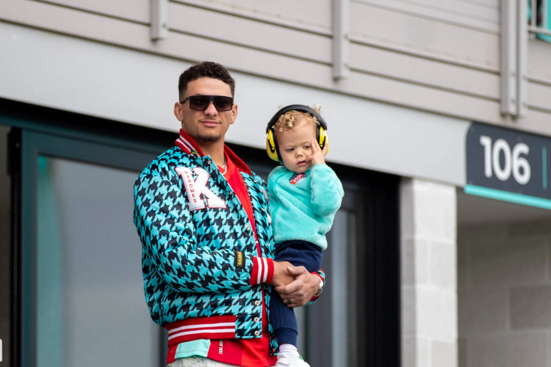 Patrick Mahomes mit Sonnenbrille, rotem Hemd und blauer Jacke mit Hahnentrittmuster hält ein kleines Kind in einem blaugrünen Pullover und gelben Kopfhörern mit Geräuschunterdrückung draußen in der Nähe von Gebäude 106". Beide schauen in die Kamera, vielleicht auf dem Weg zu einer NFL 18 Spiele-Veranstaltung. Das Kind berührt die Kopfhörer. Diese Beschreibung wurde mit der FootballR KI automatisch generiert.
