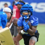 Ein Football-Spieler in blauer Ausrüstung, Helm und goldenem Visier, der Kyren Williams ähnelt, hält einen Ball, während er während des Trainings läuft. Ein Trainer in blauen und weißen Shorts hält links einen Blockierblock. Andere Spieler und ein verschwommener blauer Hintergrund sind zu sehen. Diese Beschreibung wurde mit der FootballR KI automatisch generiert.