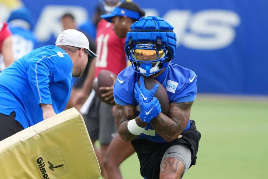 Ein Football-Spieler in blauer Ausrüstung, Helm und goldenem Visier, der Kyren Williams ähnelt, hält einen Ball, während er während des Trainings läuft. Ein Trainer in blauen und weißen Shorts hält links einen Blockierblock. Andere Spieler und ein verschwommener blauer Hintergrund sind zu sehen. Diese Beschreibung wurde mit der FootballR KI automatisch generiert.