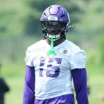 Dallas Turner steht mit lila Helm, weißem Trikot mit der Nummer 15 und lila Ärmeln auf einer Wiese und lässt sich von keinem Betrugsskandal ablenken. Ein grüner Mundschutz hängt an seinem Helm, hinter ihm verschwimmen Menschen und ein gelber Torpfosten. Diese Beschreibung wurde mit der FootballR KI automatisch generiert.