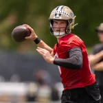 NFL Draft 2025 - Ein konzentrierter Footballspieler in einem roten Trikot und einem goldenen Helm, der Tyler Shough ähnelt, bereitet sich darauf vor, einen Football während des Trainings zu werfen. Sein Arm ist nach hinten gestreckt, während eine andere Person im unscharfen Hintergrund inmitten grüner Bäume im Freien steht. Diese Beschreibung wurde mit der FootballR KI automatisch generiert.