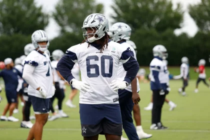 Ein Footballspieler, Tyler Guyton, der eine Uniform und einen Helm der Dallas Cowboys mit der Trikotnummer 60 trägt, steht auf einem Übungsplatz und stützt die Hände in die Hüften. Andere Spieler in ähnlicher Ausrüstung sind im Hintergrund zu sehen, zusammen mit grünen Bäumen und bewölktem Himmel. Diese Beschreibung wurde mit der FootballR KI automatisch generiert.