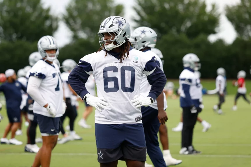 Ein Footballspieler, Tyler Guyton, der eine Uniform und einen Helm der Dallas Cowboys mit der Trikotnummer 60 trägt, steht auf einem Übungsplatz und stützt die Hände in die Hüften. Andere Spieler in ähnlicher Ausrüstung sind im Hintergrund zu sehen, zusammen mit grünen Bäumen und bewölktem Himmel. Diese Beschreibung wurde mit der FootballR KI automatisch generiert.