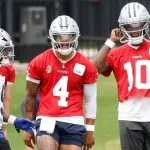 Drei Spieler der Dallas Cowboys, die zur Offense der Cowboys gehören, unterhalten sich auf dem Übungsplatz. Zwei von ihnen tragen rote Trikots und silberne Helme - die Nummer 4 - Dak Prescott - in der Mitte schreit, die Nummer 10 rückt ihren Helm zurecht - während der Spieler ganz links ein weißes Trikot mit blauen Akzenten trägt. Diese Beschreibung wurde mit der FootballR KI automatisch generiert.