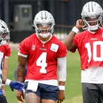 Drei Spieler der Dallas Cowboys, die zur Offense der Cowboys gehören, unterhalten sich auf dem Übungsplatz. Zwei von ihnen tragen rote Trikots und silberne Helme - die Nummer 4 - Dak Prescott - in der Mitte schreit, die Nummer 10 rückt ihren Helm zurecht - während der Spieler ganz links ein weißes Trikot mit blauen Akzenten trägt. Diese Beschreibung wurde mit der FootballR KI automatisch generiert.