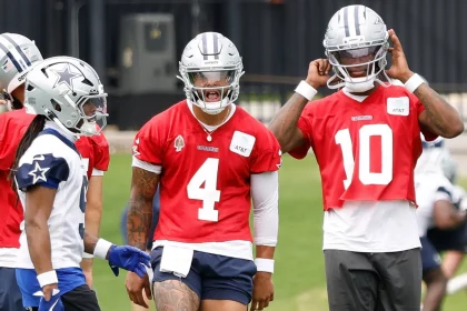 Drei Spieler der Dallas Cowboys, die zur Offense der Cowboys gehören, unterhalten sich auf dem Übungsplatz. Zwei von ihnen tragen rote Trikots und silberne Helme - die Nummer 4 - Dak Prescott - in der Mitte schreit, die Nummer 10 rückt ihren Helm zurecht - während der Spieler ganz links ein weißes Trikot mit blauen Akzenten trägt. Diese Beschreibung wurde mit der FootballR KI automatisch generiert.
