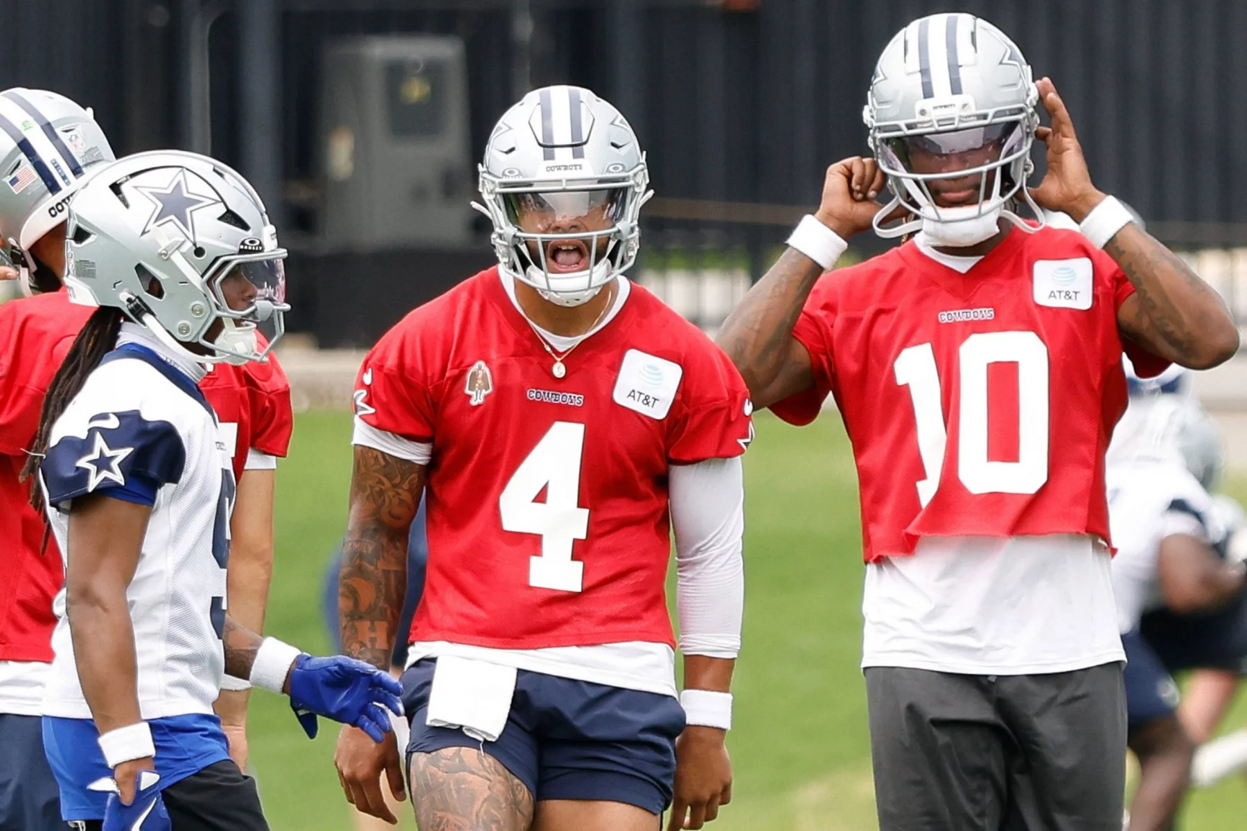 Drei Spieler der Dallas Cowboys, die zur Offense der Cowboys gehören, unterhalten sich auf dem Übungsplatz. Zwei von ihnen tragen rote Trikots und silberne Helme - die Nummer 4 - Dak Prescott - in der Mitte schreit, die Nummer 10 rückt ihren Helm zurecht - während der Spieler ganz links ein weißes Trikot mit blauen Akzenten trägt. Diese Beschreibung wurde mit der FootballR KI automatisch generiert.