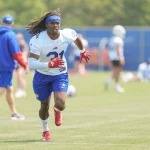 Maxwell Hairston - Ein Footballspieler in einem weißen Trikot, blauen Shorts und roten Handschuhen läuft während des Trainings auf einem grasbewachsenen Feld. Er trägt ein blaues Stirnband und Stollenschuhe. Die Szene konzentriert sich auf die Leichtathletik, ohne Hinweis auf sexuelle Gewalt, unter einem hellen Himmel. Diese Beschreibung wurde mit der FootballR KI automatisch generiert.