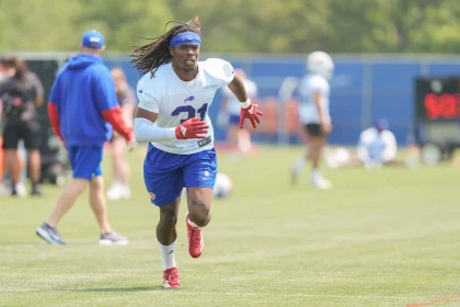 Maxwell Hairston - Ein Footballspieler in einem weißen Trikot, blauen Shorts und roten Handschuhen läuft während des Trainings auf einem grasbewachsenen Feld. Er trägt ein blaues Stirnband und Stollenschuhe. Die Szene konzentriert sich auf die Leichtathletik, ohne Hinweis auf sexuelle Gewalt, unter einem hellen Himmel. Diese Beschreibung wurde mit der FootballR KI automatisch generiert.