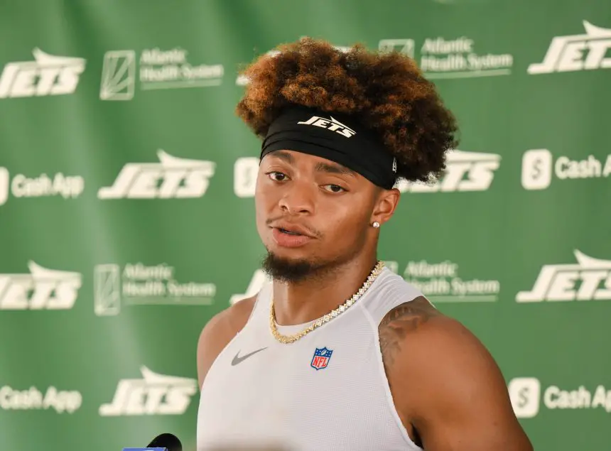 Justin Fields mit lockigem braunem Haar und einem schwarzen Jets-Stirnband spricht bei einer Presseveranstaltung, möglicherweise zu Themen wie einer Justin Fields Verletzung. Er trägt ein ärmelloses weißes Hemd, eine goldene Kette und steht vor einem Hintergrund mit grünem Jets-Sponsorenlogo. Diese Beschreibung wurde mit der FootballR KI automatisch generiert.