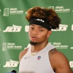 Justin Fields mit lockigem braunem Haar und einem schwarzen Jets-Stirnband spricht bei einer Presseveranstaltung, möglicherweise zu Themen wie einer Justin Fields Verletzung. Er trägt ein ärmelloses weißes Hemd, eine goldene Kette und steht vor einem Hintergrund mit grünem Jets-Sponsorenlogo. Diese Beschreibung wurde mit der FootballR KI automatisch generiert.