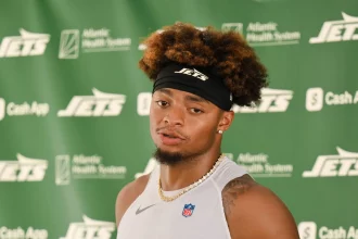 Justin Fields mit lockigem braunem Haar und einem schwarzen Jets-Stirnband spricht bei einer Presseveranstaltung, möglicherweise zu Themen wie einer Justin Fields Verletzung. Er trägt ein ärmelloses weißes Hemd, eine goldene Kette und steht vor einem Hintergrund mit grünem Jets-Sponsorenlogo. Diese Beschreibung wurde mit der FootballR KI automatisch generiert.