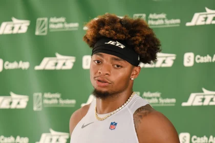 Justin Fields mit lockigem braunem Haar und einem schwarzen Jets-Stirnband spricht bei einer Presseveranstaltung, möglicherweise zu Themen wie einer Justin Fields Verletzung. Er trägt ein ärmelloses weißes Hemd, eine goldene Kette und steht vor einem Hintergrund mit grünem Jets-Sponsorenlogo. Diese Beschreibung wurde mit der FootballR KI automatisch generiert.