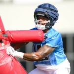 Treylon Burks, der ein blaues Trikot und einen gepolsterten Helm trägt, schiebt beim Training einen roten Tackling-Dummy. Er trägt weiße Handschuhe und sein Blick ist konzentriert. Der Hintergrund ist unscharf, um die Action und Intensität der Übung zu unterstreichen. Diese Beschreibung wurde mit der FootballR KI automatisch generiert.