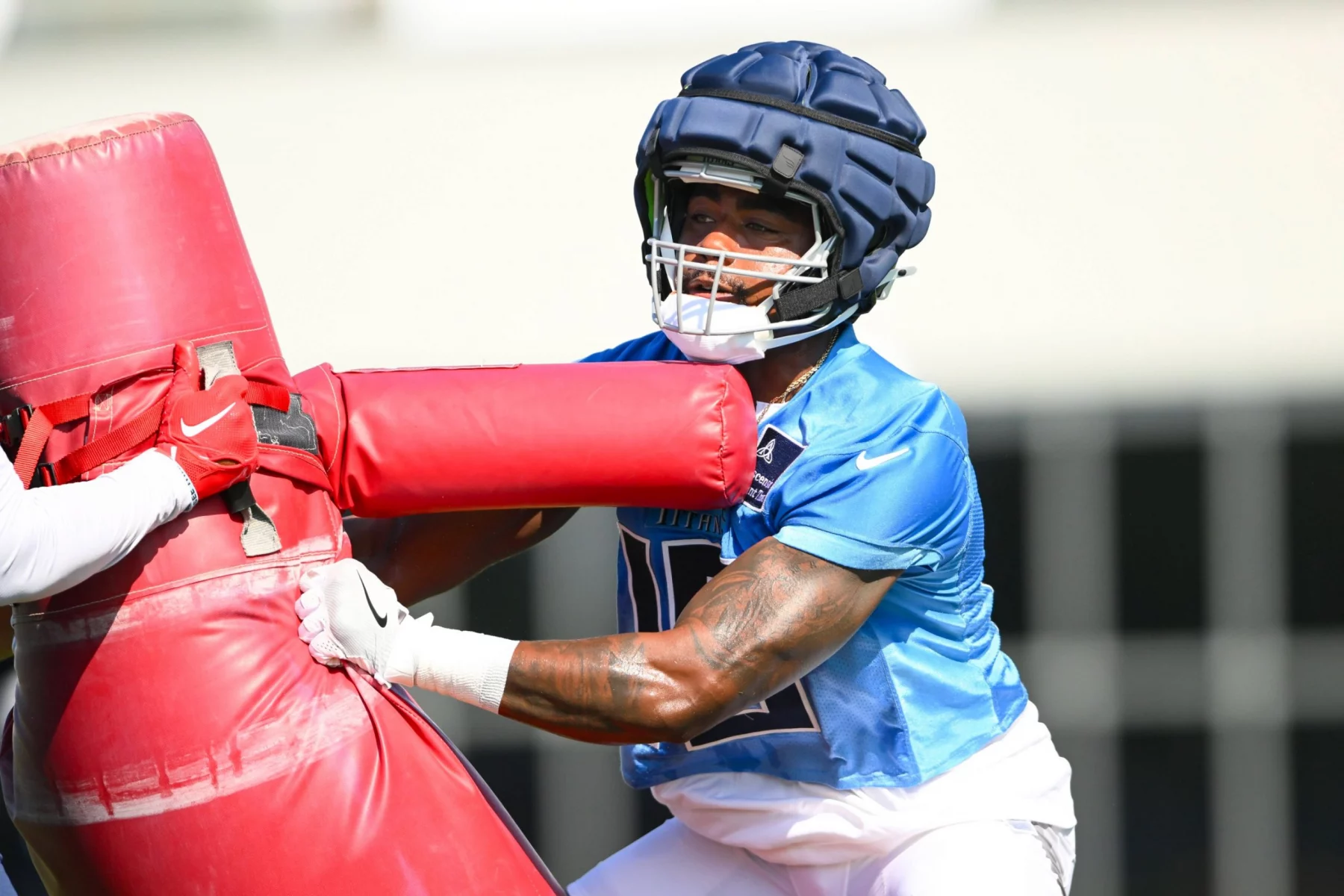 Treylon Burks, der ein blaues Trikot und einen gepolsterten Helm trägt, schiebt beim Training einen roten Tackling-Dummy. Er trägt weiße Handschuhe und sein Blick ist konzentriert. Der Hintergrund ist unscharf, um die Action und Intensität der Übung zu unterstreichen. Diese Beschreibung wurde mit der FootballR KI automatisch generiert.