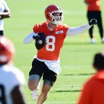 Roster Cuts - Ein Footballspieler in einem orangefarbenen Trikot mit der Nummer 8, das an Kenny Pickett erinnert, und einem weißen Helm läuft auf einem Feld, hält einen Football in der rechten Hand und zeigt mit der linken auf ihn. Andere Spieler in orangefarbenen und weißen Trainingsanzügen sind in der Nähe zu sehen. Diese Beschreibung wurde mit der FootballR KI automatisch generiert.