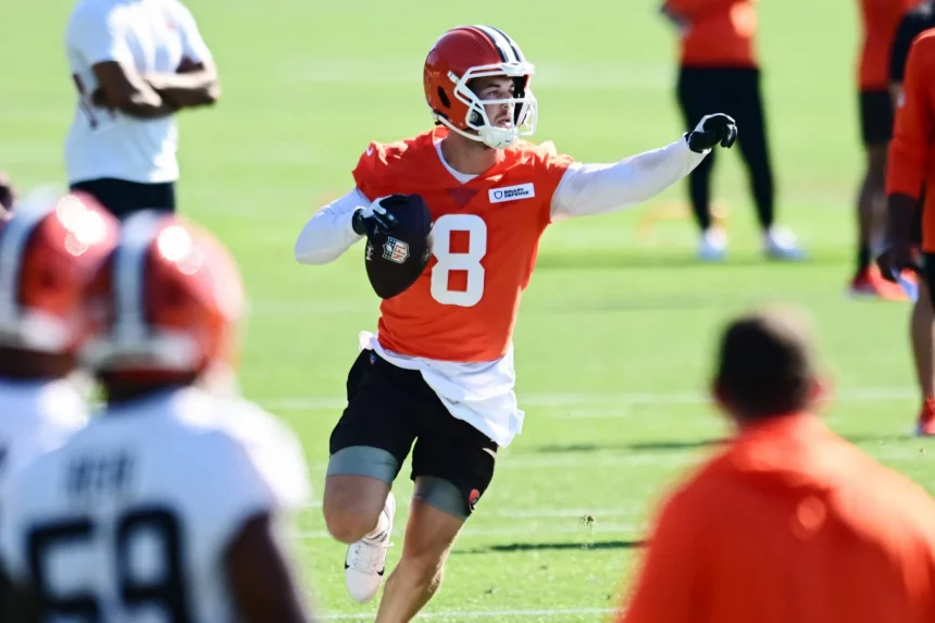 Roster Cuts - Ein Footballspieler in einem orangefarbenen Trikot mit der Nummer 8, das an Kenny Pickett erinnert, und einem weißen Helm läuft auf einem Feld, hält einen Football in der rechten Hand und zeigt mit der linken auf ihn. Andere Spieler in orangefarbenen und weißen Trainingsanzügen sind in der Nähe zu sehen. Diese Beschreibung wurde mit der FootballR KI automatisch generiert.