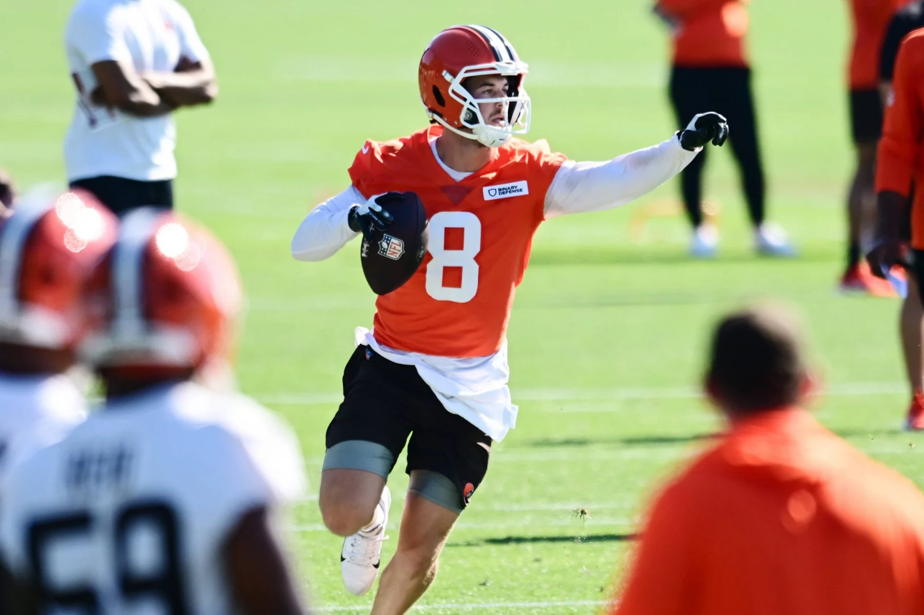 Roster Cuts - Ein Footballspieler in einem orangefarbenen Trikot mit der Nummer 8, das an Kenny Pickett erinnert, und einem weißen Helm läuft auf einem Feld, hält einen Football in der rechten Hand und zeigt mit der linken auf ihn. Andere Spieler in orangefarbenen und weißen Trainingsanzügen sind in der Nähe zu sehen. Diese Beschreibung wurde mit der FootballR KI automatisch generiert.