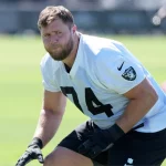 Kolton Miller, ein männlicher Football-Spieler in einem weißen Las Vegas Raiders-Trikot mit der Nummer 74, trainiert im Freien auf einem grünen Feld. Er trägt schwarze Handschuhe und hockt in einer athletischen Haltung. Er schaut konzentriert zwischen anderen Spielern und verschwommenen Feldelementen. Diese Beschreibung wurde mit der FootballR KI automatisch generiert.