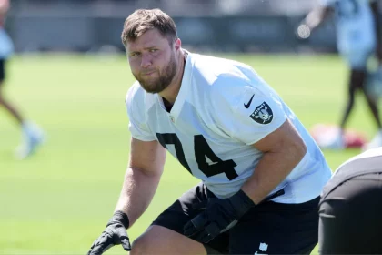 Kolton Miller, ein männlicher Football-Spieler in einem weißen Las Vegas Raiders-Trikot mit der Nummer 74, trainiert im Freien auf einem grünen Feld. Er trägt schwarze Handschuhe und hockt in einer athletischen Haltung. Er schaut konzentriert zwischen anderen Spielern und verschwommenen Feldelementen. Diese Beschreibung wurde mit der FootballR KI automatisch generiert.