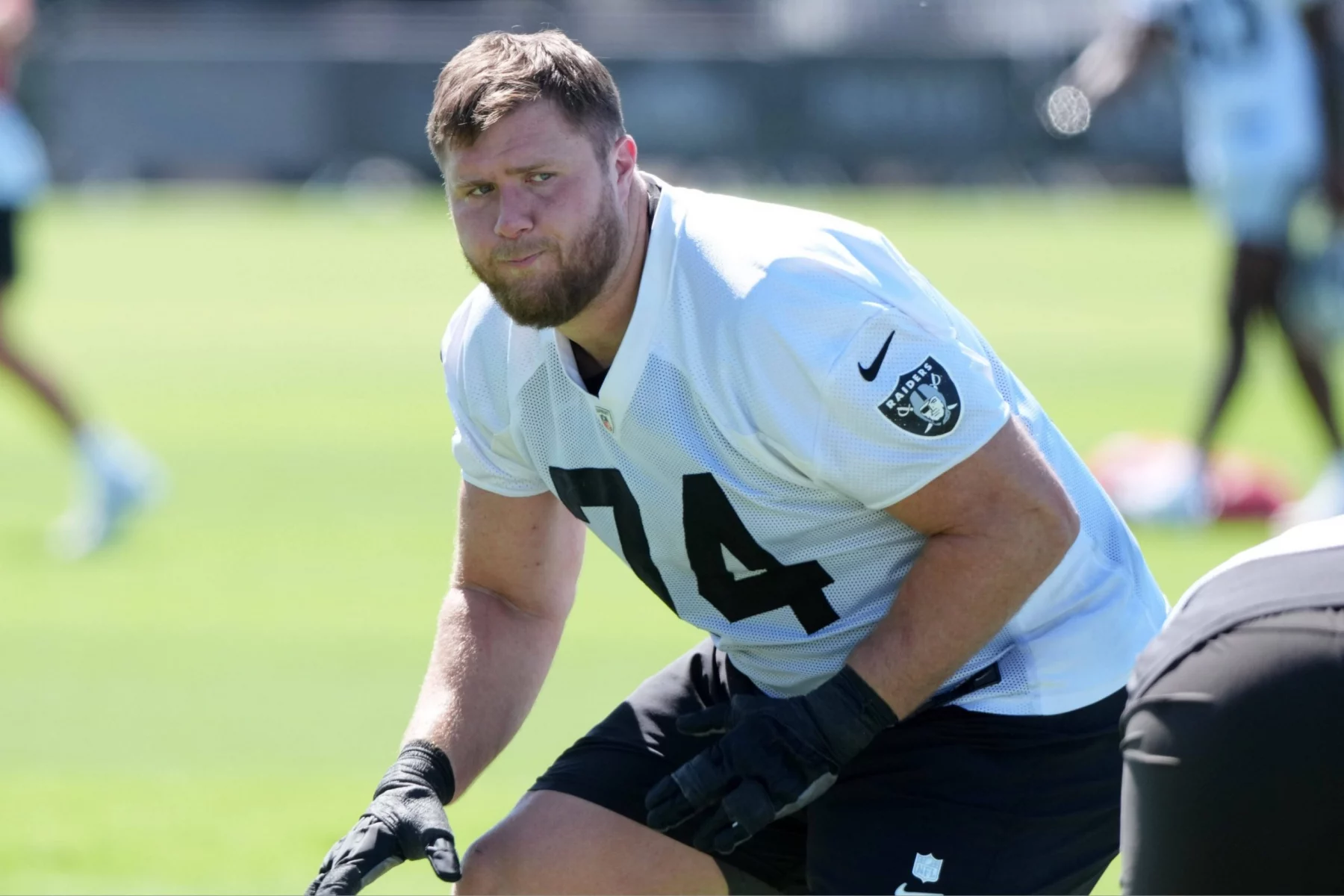 Kolton Miller, ein männlicher Football-Spieler in einem weißen Las Vegas Raiders-Trikot mit der Nummer 74, trainiert im Freien auf einem grünen Feld. Er trägt schwarze Handschuhe und hockt in einer athletischen Haltung. Er schaut konzentriert zwischen anderen Spielern und verschwommenen Feldelementen. Diese Beschreibung wurde mit der FootballR KI automatisch generiert.