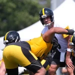 Aaron Rodgers, in weißem Trikot und Helm, steht hinter dem Center und ist bereit für den Snap während des Trainings. Teamkollegen der Steelers in gelben und schwarzen Uniformen stellen sich auf einem sonnigen Feld auf, mit einem weißen Zelt und grünen Bäumen im Hintergrund. Diese Beschreibung wurde mit der FootballR KI automatisch generiert.