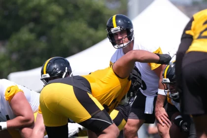 Aaron Rodgers, in weißem Trikot und Helm, steht hinter dem Center und ist bereit für den Snap während des Trainings. Teamkollegen der Steelers in gelben und schwarzen Uniformen stellen sich auf einem sonnigen Feld auf, mit einem weißen Zelt und grünen Bäumen im Hintergrund. Diese Beschreibung wurde mit der FootballR KI automatisch generiert.