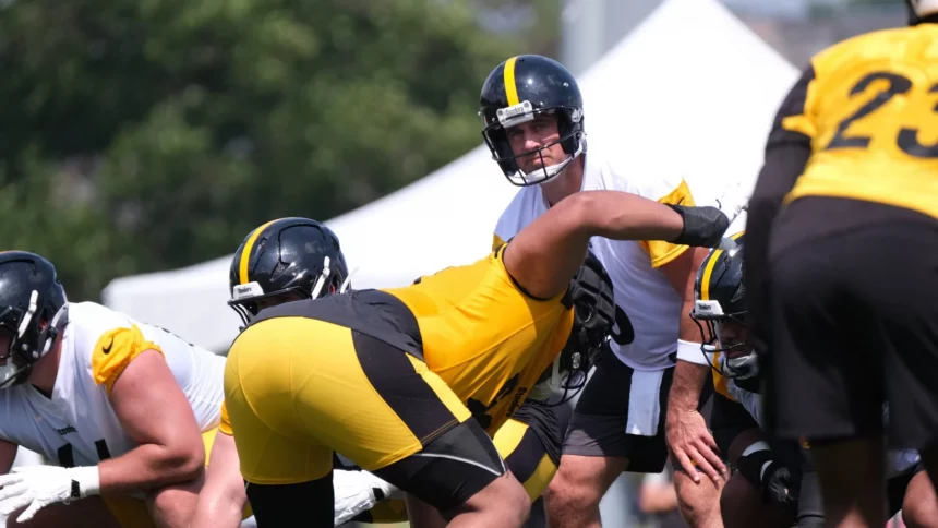 Aaron Rodgers, in weißem Trikot und Helm, steht hinter dem Center und ist bereit für den Snap während des Trainings. Teamkollegen der Steelers in gelben und schwarzen Uniformen stellen sich auf einem sonnigen Feld auf, mit einem weißen Zelt und grünen Bäumen im Hintergrund. Diese Beschreibung wurde mit der FootballR KI automatisch generiert.
