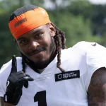 Ein lächelnder Footballspieler, Martin Emerson Jr., mit langen Dreadlocks trägt ein weißes Trikot mit "1" und "BINARY DEFENSE", einen schwarzen Handschuh und ein orangefarbenes Stirnband mit der Aufschrift "BROWNS". Er zeigt nach oben und steht im Freien mit Bäumen im Hintergrund. Diese Beschreibung wurde mit der FootballR KI automatisch generiert.