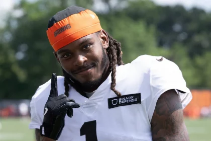 Ein lächelnder Footballspieler, Martin Emerson Jr., mit langen Dreadlocks trägt ein weißes Trikot mit "1" und "BINARY DEFENSE", einen schwarzen Handschuh und ein orangefarbenes Stirnband mit der Aufschrift "BROWNS". Er zeigt nach oben und steht im Freien mit Bäumen im Hintergrund. Diese Beschreibung wurde mit der FootballR KI automatisch generiert.