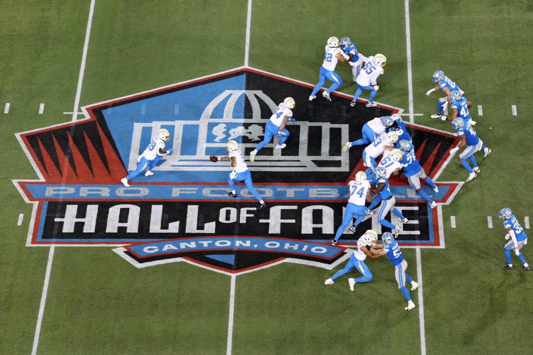 Hall of Fame Game - Luftaufnahme eines NFL Preseason Footballspiels mit Spielern in blau-weißen Uniformen auf dem Spielfeld. Der Ball wird über das große "Pro Football Hall of Fame, Canton, Ohio"-Logo in der Mitte des Spielfelds gespielt, wobei die Aktion und der ikonische Moment festgehalten werden. Diese Beschreibung wurde mit der FootballR KI automatisch generiert.