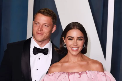 Christian McCaffrey in einem schwarzen Smoking mit Fliege steht lächelnd neben Olivia Culpo in einem schulterfreien rosa Kleid. Sie erscheinen auf einer formellen Veranstaltung mit einem stilvollen dunkel-weiß gemusterten Hintergrund hinter ihnen. Diese Beschreibung wurde mit der FootballR KI automatisch generiert.