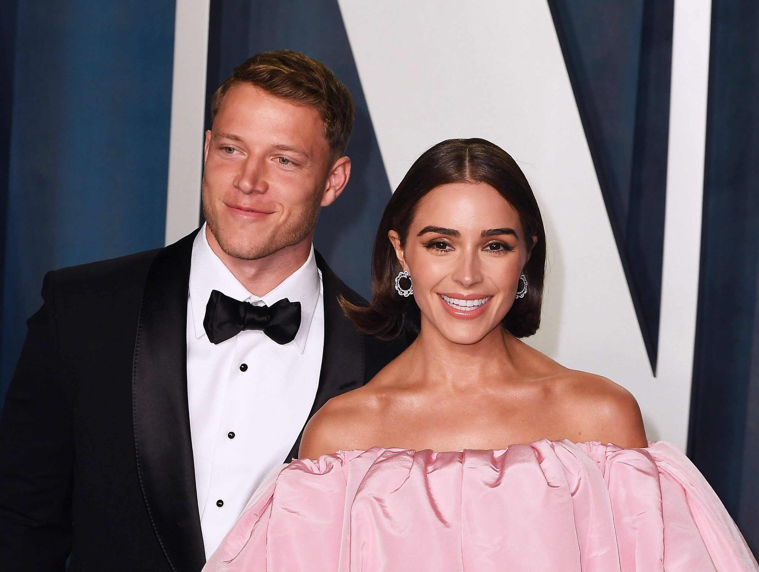 Christian McCaffrey in einem schwarzen Smoking mit Fliege steht lächelnd neben Olivia Culpo in einem schulterfreien rosa Kleid. Sie erscheinen auf einer formellen Veranstaltung mit einem stilvollen dunkel-weiß gemusterten Hintergrund hinter ihnen. Diese Beschreibung wurde mit der FootballR KI automatisch generiert.