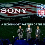 Drei Männer in Anzügen stehen auf einer Bühne vor großen SONY- und NFL-Logos, auf denen "A TECHNOLOGY PARTNER OF THE NFL" steht. Die Zuschauer halten Telefone für Fotos hoch, während Sony Headsets als wichtiges Hilfsmittel für NFL-Trainer in der Saison 2025 hervorgehoben werden. Diese Beschreibung wurde mit der FootballR KI automatisch generiert.