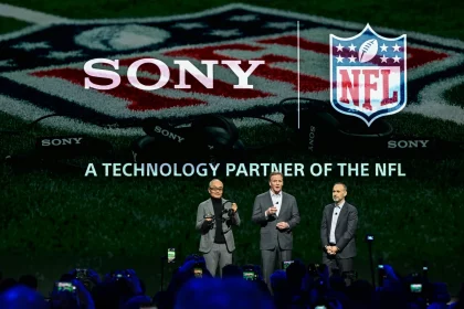 Drei Männer in Anzügen stehen auf einer Bühne vor großen SONY- und NFL-Logos, auf denen "A TECHNOLOGY PARTNER OF THE NFL" steht. Die Zuschauer halten Telefone für Fotos hoch, während Sony Headsets als wichtiges Hilfsmittel für NFL-Trainer in der Saison 2025 hervorgehoben werden. Diese Beschreibung wurde mit der FootballR KI automatisch generiert.